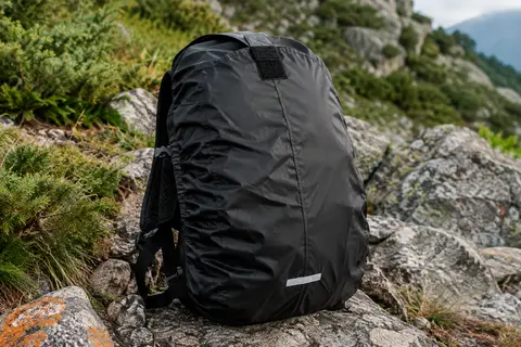 Housse de pluie Eastpak Cory noire en polyester, posée sur un sac à dos sur un sol minéral de montagne.