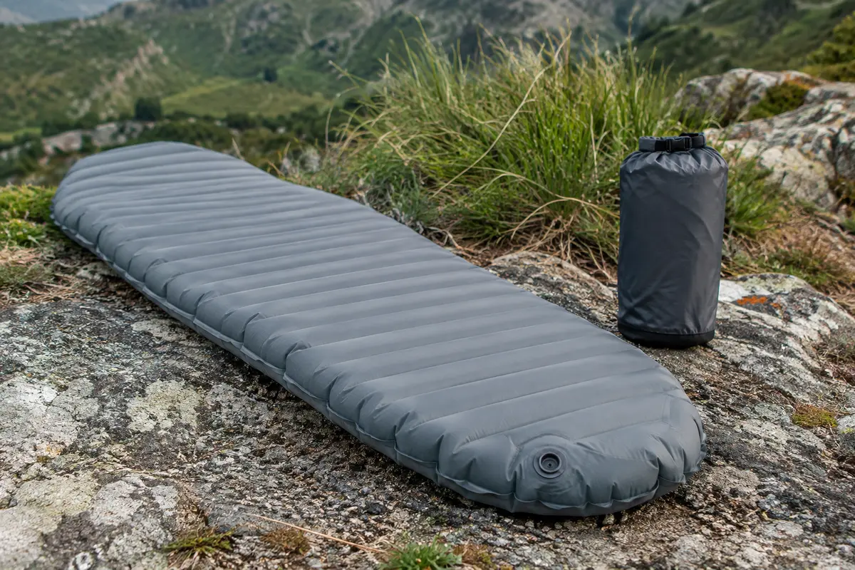 Matelas de randonnée gonflable gris en forme momie, posé sur un sol minéral près de son sac de pompe.