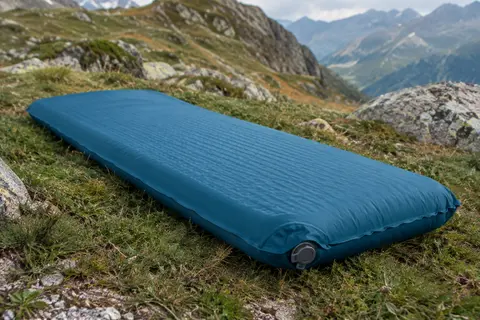 Matelas gonflable de randonnée bleu paon déployé sur une herbe de montagne près d’un sol minéral