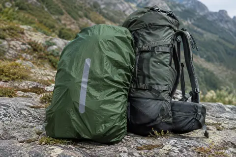 Housse de pluie verte pour sac à dos, avec bande réfléchissante et sangle extensible, posée sur un sol minéral.