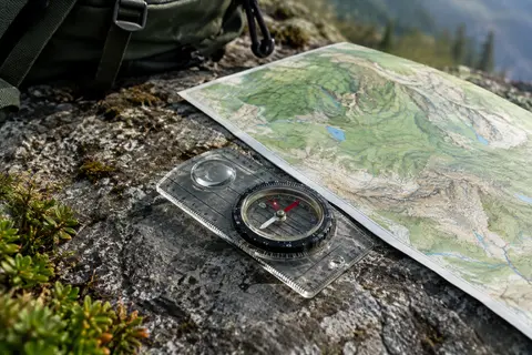 Boussole à base transparente, lunette rotative et loupe posée sur une carte sans inscriptions, sur un sol minéral.