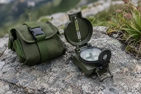 Boussole lensatique vert armée avec son étui, posée sur une pierre grise en montagne.