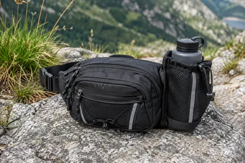 Sac banane de randonnée en nylon noir avec porte-bouteille, posé sur un sol minéral en montagne.