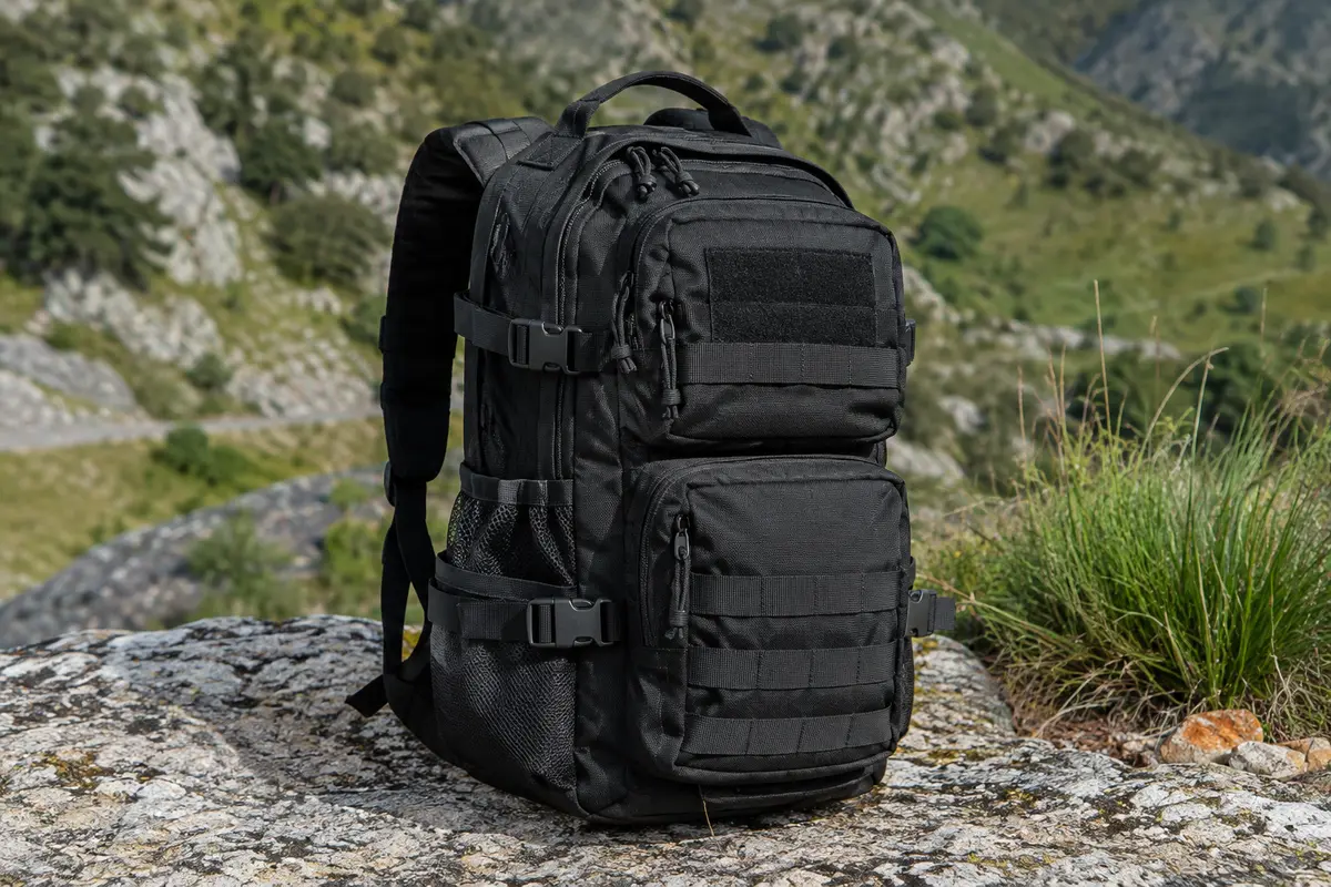Sac à dos tactique noir de 30 L avec compartiments et poches latérales, posé sur un sol minéral